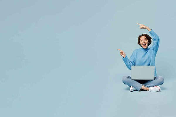 Eine Frau sitzt im Schneidersitz auf dem Boden, auf Ihren Beinen steht ein Laptop.